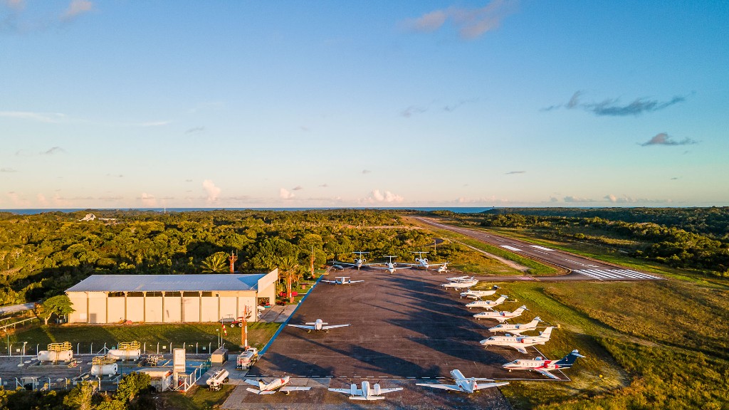 Vista aérea do aeródromo com pátio, várias aeronaves estacionadas e mata ao redor ao entardecer.